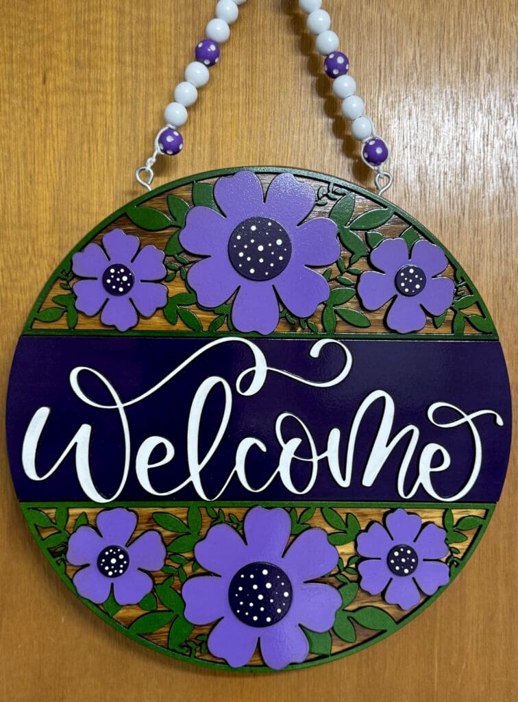 Round welcome sign with purple flowers.